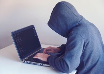 A person wearing a grey hoodie with the hood pulled up sits at a white desk, typing on a laptop displaying lines of computer code. The image represents cybercriminal activity, such as credential stuffing and account takeover fraud, by keeping the user's identity hidden.