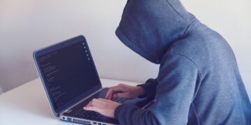 A person wearing a grey hoodie with the hood pulled up sits at a white desk, typing on a laptop displaying lines of computer code. The image represents cybercriminal activity, such as credential stuffing and account takeover fraud, by keeping the user's identity hidden.
