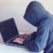 A person wearing a grey hoodie with the hood pulled up sits at a white desk, typing on a laptop displaying lines of computer code. The image represents cybercriminal activity, such as credential stuffing and account takeover fraud, by keeping the user's identity hidden.