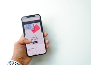 A close-up, first-person perspective of a hand holding a smartphone against a clean white background. The phone screen displays an advertisement for Airbnb gift cards with a "Shop now" button, representing a target for digital asset and loyalty fraud schemes.