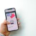 A close-up, first-person perspective of a hand holding a smartphone against a clean white background. The phone screen displays an advertisement for Airbnb gift cards with a "Shop now" button, representing a target for digital asset and loyalty fraud schemes.
