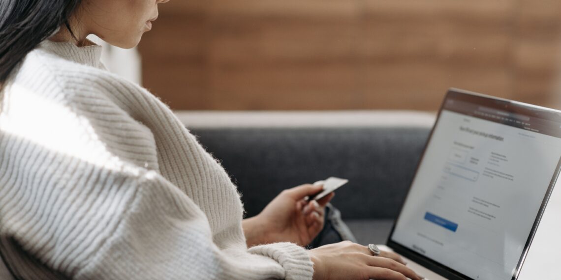 A woman sitting on a sofa while typing on her laptop and holding a credit card, illustrating the secure online login and verification processes discussed in "What Is Multi-Factor Authentication?".