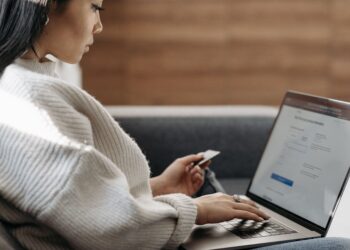 A woman sitting on a sofa while typing on her laptop and holding a credit card, illustrating the secure online login and verification processes discussed in "What Is Multi-Factor Authentication?".