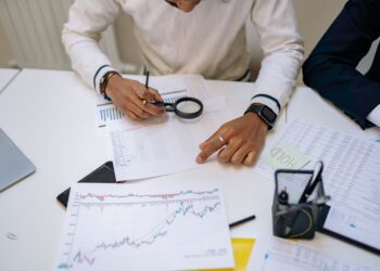 Fraud analyst using a magnifying glass to review financial charts and transaction data on a desk with a laptop and stock reports.