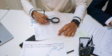 Fraud analyst using a magnifying glass to review financial charts and transaction data on a desk with a laptop and stock reports.