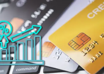 Close-up shot of multiple credit cards scattered over a black laptop keyboard. A prominent gold credit card sits in the foreground. Layered on top of the left side is a bright cyan line icon showing a rising bar graph with a percentage sign and an upward-pointing arrow, symbolizing an increase in rates or credit card fraud statistics.