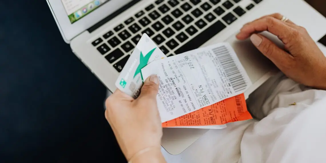 A person seated with a laptop reconciles their spending, holding a debit card alongside printed receipts while checking transaction records on screen — an everyday moment of expense-tracking that can also help spot signs of first-party fraud, such as disputing legitimate purchases.