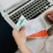 A person seated with a laptop reconciles their spending, holding a debit card alongside printed receipts while checking transaction records on screen — an everyday moment of expense-tracking that can also help spot signs of first-party fraud, such as disputing legitimate purchases.