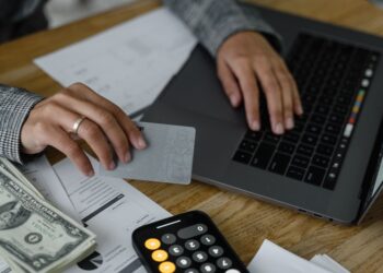 Close-up of a person's hands at a desk, one holding a gray card while the other types on a laptop keyboard, with cash, a calculator, and papers labeled "Expense Type" and "Revenue by Division" spread across the table.