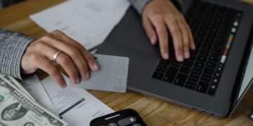 Close-up of a person's hands at a desk, one holding a gray card while the other types on a laptop keyboard, with cash, a calculator, and papers labeled "Expense Type" and "Revenue by Division" spread across the table.