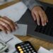 Close-up of a person's hands at a desk, one holding a gray card while the other types on a laptop keyboard, with cash, a calculator, and papers labeled "Expense Type" and "Revenue by Division" spread across the table.