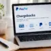 A laptop on a wooden desk displays a webpage titled "PayPal Chargebacks," explaining that a chargeback is a refund request made by a cardholder through their bank or card issuer, with a flow diagram showing the process moving from Cardholder to Issuing Bank to PayPal to Merchant. A coffee mug, potted plant, notebook, and pen sit nearby on the desk.