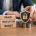 Wooden blocks reading 'Fraud Prevention Budget' next to a hand placing a padlock shield on stacks of coins.