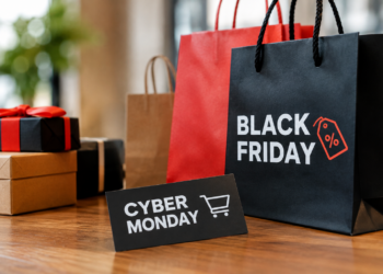 Stock photo of shopping bags labeled “Black Friday” and a “Cyber Monday” card with a shopping cart icon, beside wrapped gift boxes on a wooden table, illustrating holiday shopping sales.
