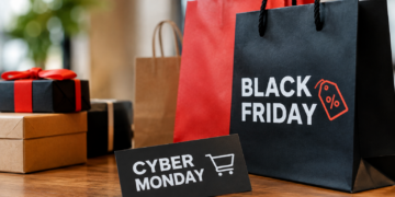 Stock photo of shopping bags labeled “Black Friday” and a “Cyber Monday” card with a shopping cart icon, beside wrapped gift boxes on a wooden table, illustrating holiday shopping sales.