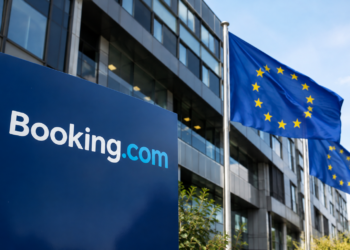 Stock photo of a blue “Booking.com” sign outside an office building, with European Union flags flying nearby against a clear sky.