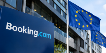 Stock photo of a blue “Booking.com” sign outside an office building, with European Union flags flying nearby against a clear sky.