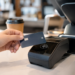 Stock photo of a hand tapping a contactless credit card against a payment terminal at a café counter, with a coffee cup and pastry display in the background.