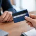 Close-up of a hand holding the back of a blue credit card with the three-digit Card Verification Value (CVV) printed beside the signature strip, held out across a wooden desk toward another person, with a laptop and spiral notebook in the foreground.