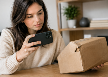 Stock photo of a woman holding a smartphone to photograph or scan an empty, damaged cardboard box at a table, illustrating documentation of a missing or damaged package delivery.