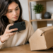 Stock photo of a woman holding a smartphone to photograph or scan an empty, damaged cardboard box at a table, illustrating documentation of a missing or damaged package delivery.