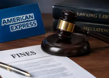 Stock photo of a judge’s gavel resting beside a document titled “Fines,” with an American Express sign and law books labeled “Banking Law” and “Regulations” in the background, illustrating regulatory fines or legal action against a financial company.