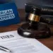 Stock photo of a judge’s gavel resting beside a document titled “Fines,” with an American Express sign and law books labeled “Banking Law” and “Regulations” in the background, illustrating regulatory fines or legal action against a financial company.