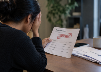 Stock photo of a distressed woman holding her head while reading a bank statement marked “Fraud Alert,” listing several pending online and card-not-present charges, with additional mail and an “Account Alert” envelope on the desk beside her.