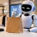 Stock photo of a white humanoid robot with glowing blue eyes holding a brown paper shopping bag on a counter inside a clothing store, with a shopping cart icon displayed on a screen in the background, illustrating AI-assisted retail shopping.
