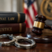Stock photo of a judge’s gavel and handcuffs on a wooden desk beside books titled “Criminal Law” and “Justice,” with an American flag and an official seal in the background.