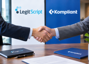 Stock photo of two businesspeople shaking hands over a desk, with LegitScript and Kompliant branded folders, a pen, and a signed document on the table, and company logo banners in the background, symbolizing a partnership between the two compliance companies.