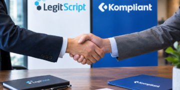 Stock photo of two businesspeople shaking hands over a desk, with LegitScript and Kompliant branded folders, a pen, and a signed document on the table, and company logo banners in the background, symbolizing a partnership between the two compliance companies.
