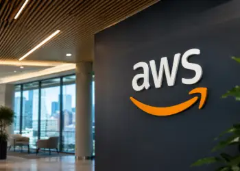 Stock photo of the AWS logo displayed in 3D lettering on a dark wall inside a modern office lobby, with a city skyline visible through floor-to-ceiling windows and seating in the background.