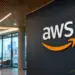Stock photo of the AWS logo displayed in 3D lettering on a dark wall inside a modern office lobby, with a city skyline visible through floor-to-ceiling windows and seating in the background.