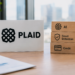 Stock photo of an office desk with a white nameplate displaying the Plaid logo beside a stack of three wooden blocks labeled "AI," "Fraud Detection," and "Credit," each with a matching icon, evoking AI-driven fraud detection in financial data and credit services.