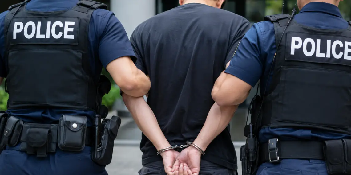 Stock photo of a handcuffed man being escorted by two police officers in tactical vests, viewed from behind, outside a building.