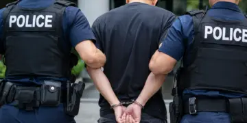 Stock photo of a handcuffed man being escorted by two police officers in tactical vests, viewed from behind, outside a building.