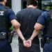 Stock photo of a handcuffed man being escorted by two police officers in tactical vests, viewed from behind, outside a building.