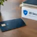 Dark blue contactless chip card lying on a wooden desk beside a white sign with a blue shield and checkmark icon reading “3D Secure: Safer Payments. Greater Confidence,” with a potted plant and stacked notebooks in the background.