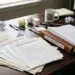 Messy desk piled with compliance notices, surcharging regulation updates, and a merchant processing agreement, beside an ISV compliance manual, a calendar marked with new rule dates, and a 'Small Business Owner' mug.