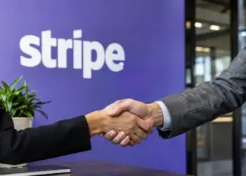 Two people in business attire shake hands in front of a purple wall with the Stripe logo.