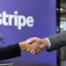 Two people in business attire shake hands in front of a purple wall with the Stripe logo.