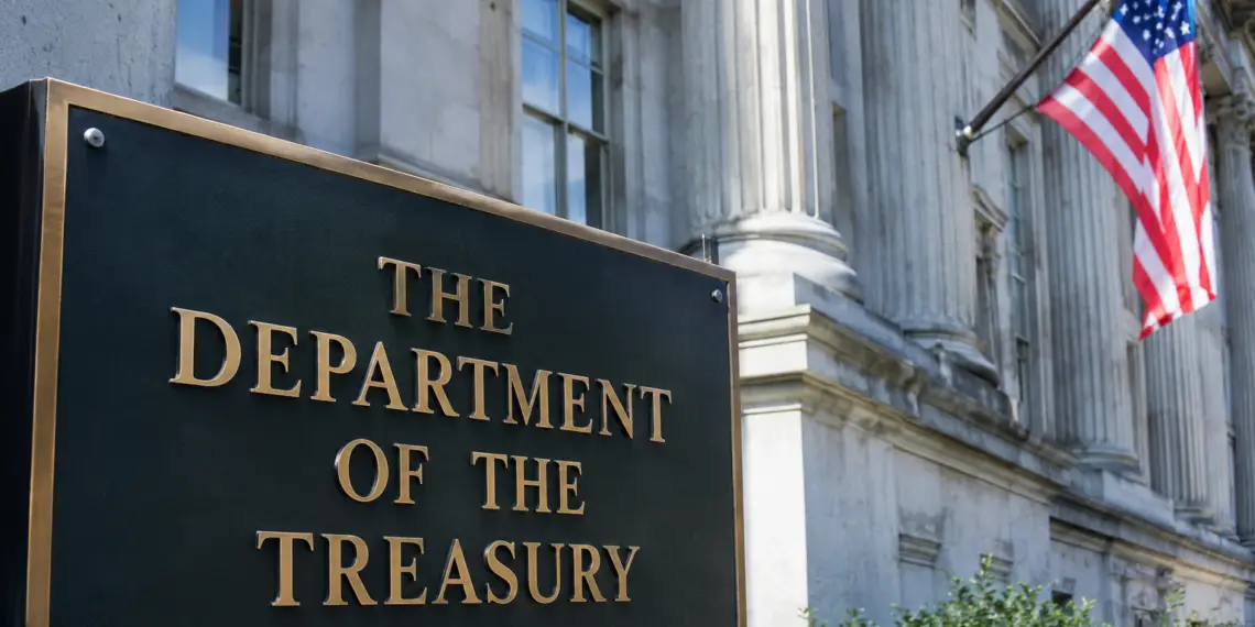 Stock photo of the bronze “The Department of the Treasury” sign outside the U.S. Treasury Building, with its columned facade and an American flag visible in the background.
