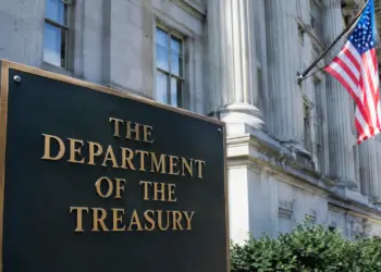 Stock photo of the bronze “The Department of the Treasury” sign outside the U.S. Treasury Building, with its columned facade and an American flag visible in the background.