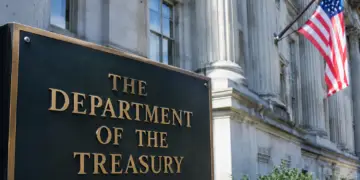 Stock photo of the bronze “The Department of the Treasury” sign outside the U.S. Treasury Building, with its columned facade and an American flag visible in the background.