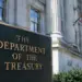 Stock photo of the bronze “The Department of the Treasury” sign outside the U.S. Treasury Building, with its columned facade and an American flag visible in the background.