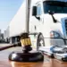 Stock photo of a judge’s gavel resting on a desk beside a pair of handcuffs on a document folder, with a semi-truck and trailers parked in the background, illustrating legal action or enforcement related to the trucking or freight industry.