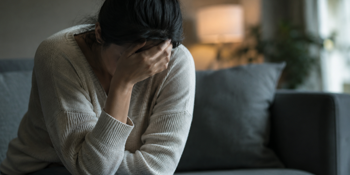 Stock photo of a woman sitting on a couch with her head resting in her hand, appearing distressed, in a dimly lit living room.