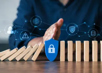 A hand stops a row of falling wooden dominoes with a blue shield bearing a padlock icon, protecting the upright dominoes beyond it. Floating digital icons above show a hooded hacker, a warning sign, a brain, a shield, and a database, connected by network lines.