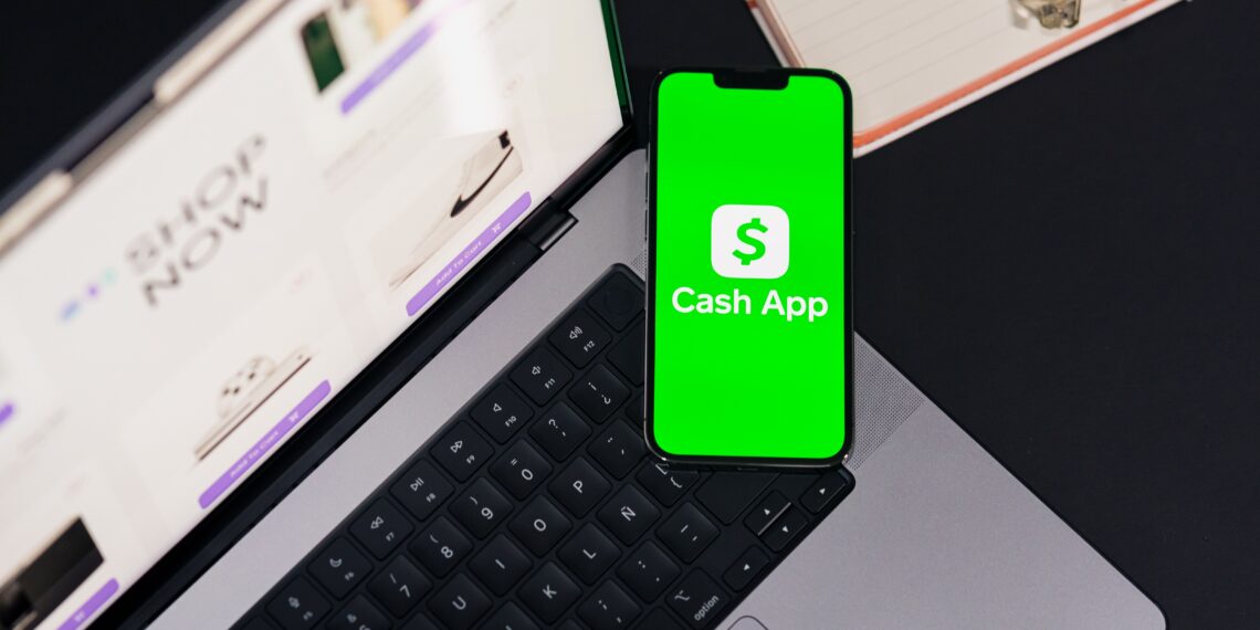 Stock photo of a smartphone displaying the Cash App logo on a green screen, resting on a laptop keyboard with an online shopping page open in the background, alongside a notebook, pen, and glasses.